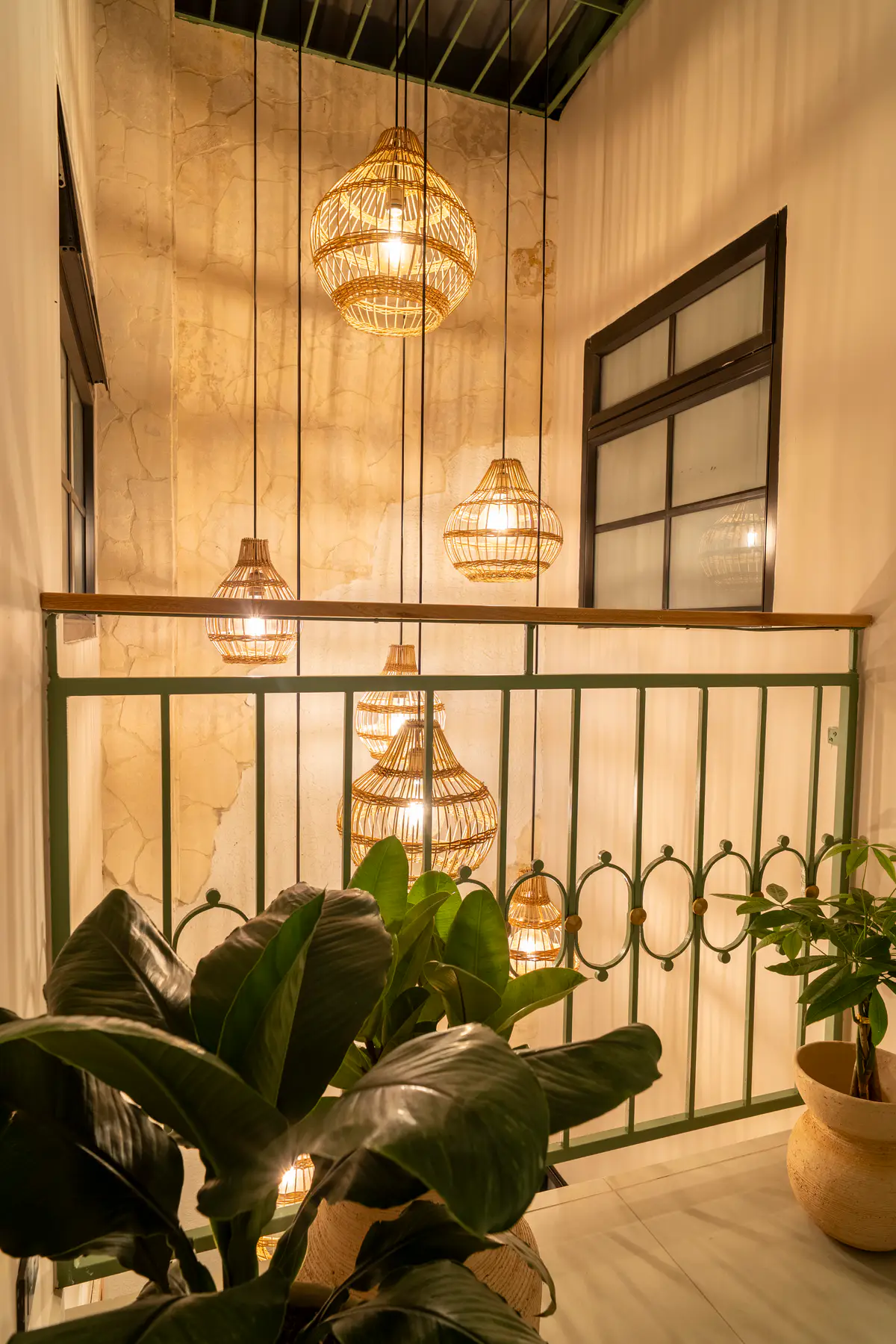 El vacío de la escalera con lámparas tejidas encendidas a distintas alturas, una baranda verde y plantas en primer plano.