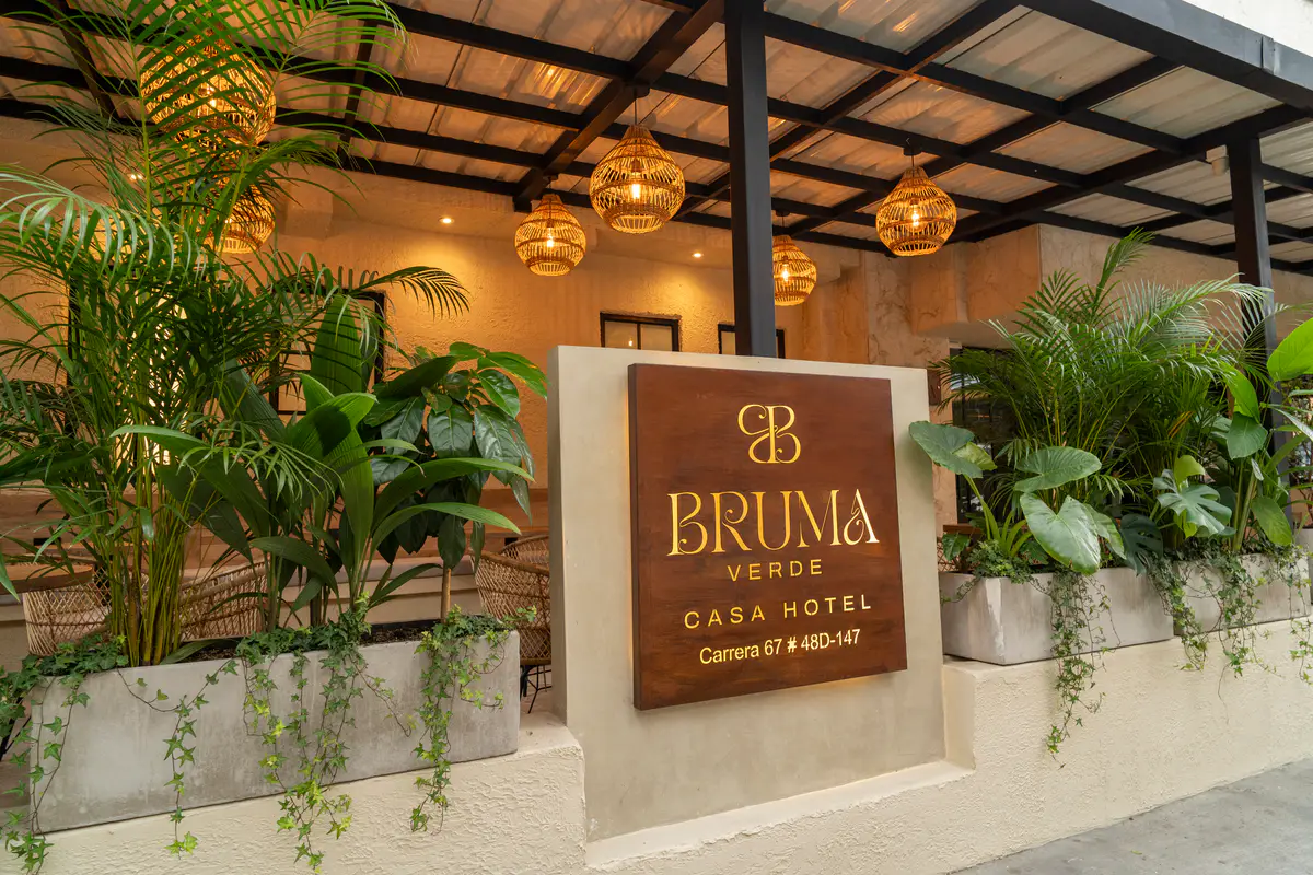 Entrada de Bruma Verde de día: letrero de madera con el monograma y la dirección Carrera 67 # 48D-147, jardineras con palmas y lámparas tejidas bajo la pérgola.