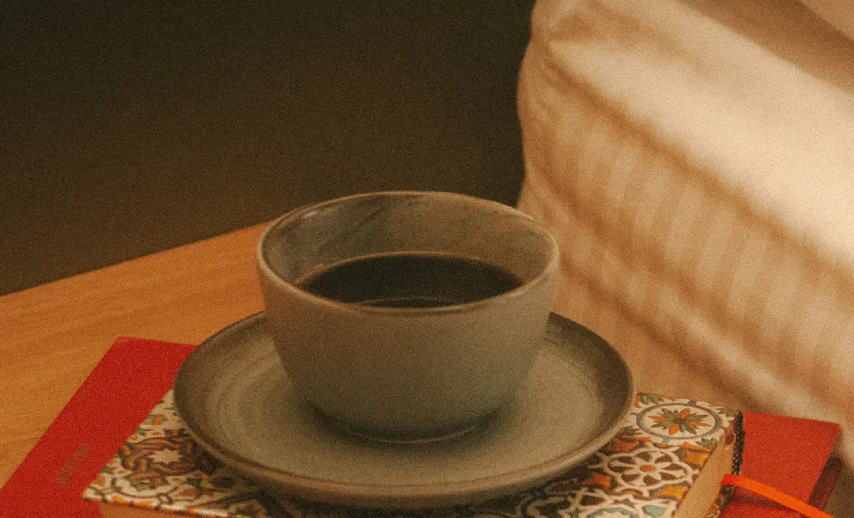 Una taza de café sobre una pila de libros, junto a la cama, en luz cálida.