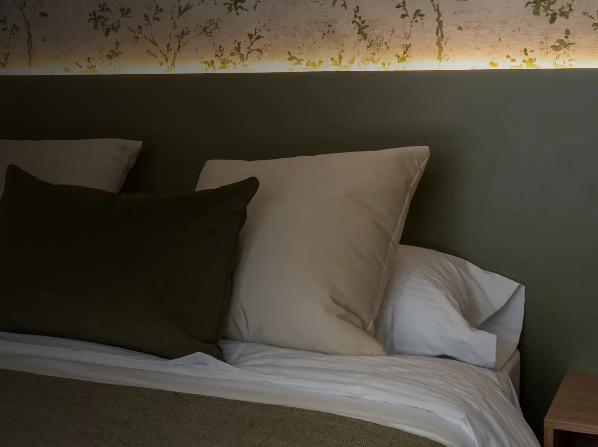 Cama tendida con cojines blancos y verde oliva, cabecero verde y papel de colgadura botánico.