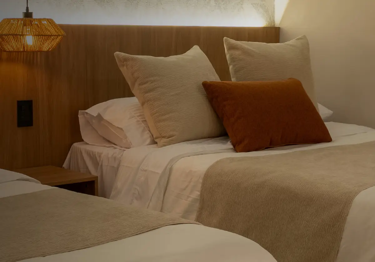 Dos camas con cojines beige y terracota, cabecero de madera y una lámpara tejida colgante.