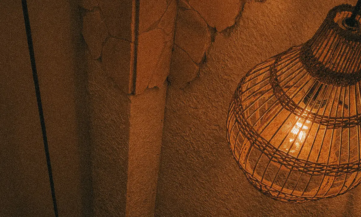 Una lámpara tejida encendida contra una pared de textura, en luz ámbar.