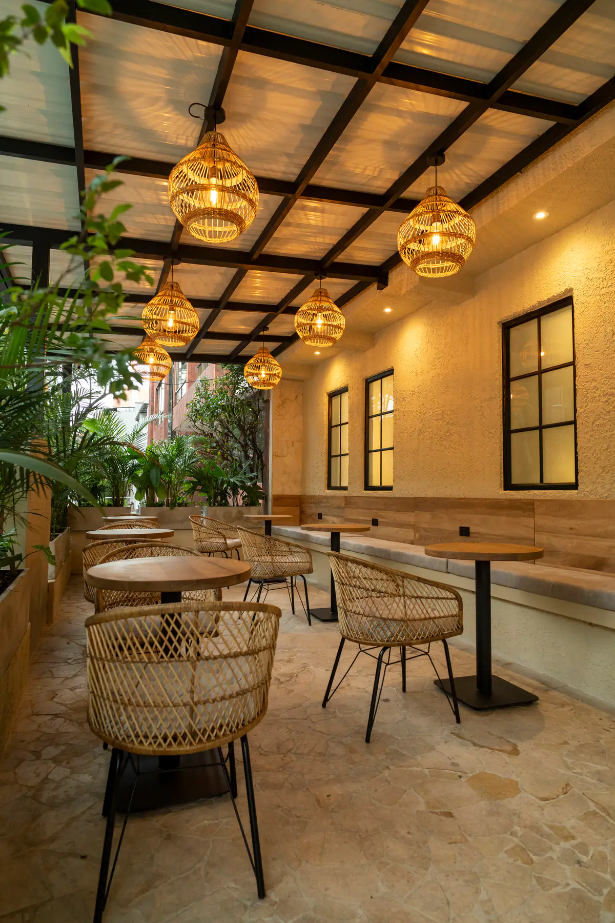 Terraza bajo la pérgola con sillas de mimbre, una banca de madera a lo largo de la pared, plantas y lámparas tejidas.